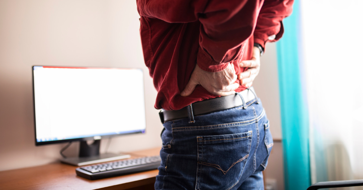 man stands in front of computer desk holding a sore lower back